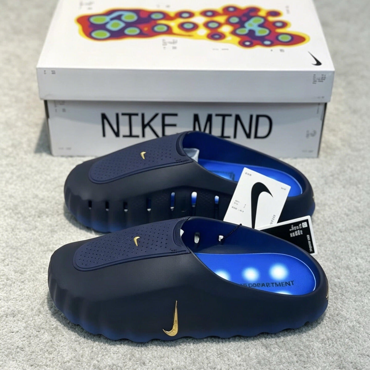 A pair of Nike Mind slip-on clogs in navy blue with gold Swoosh logos and bright blue interiors, displayed in front of their white Nike Mind retail box on a gray carpet.